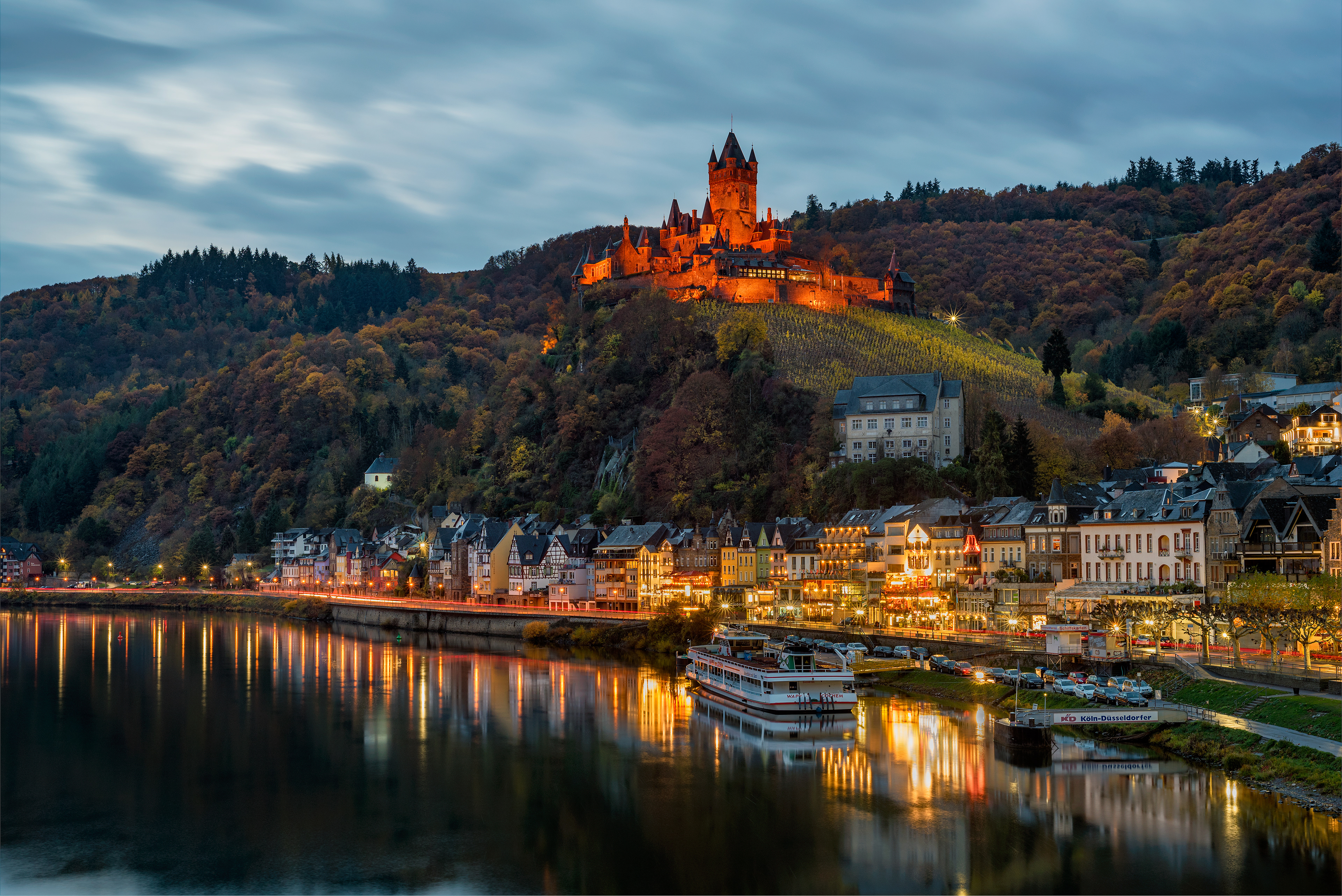 The Imperial Castle of Cochem (Reichsburg Cochem) perched high on a hill, glowing golden above the Moselle River and town promenade at night