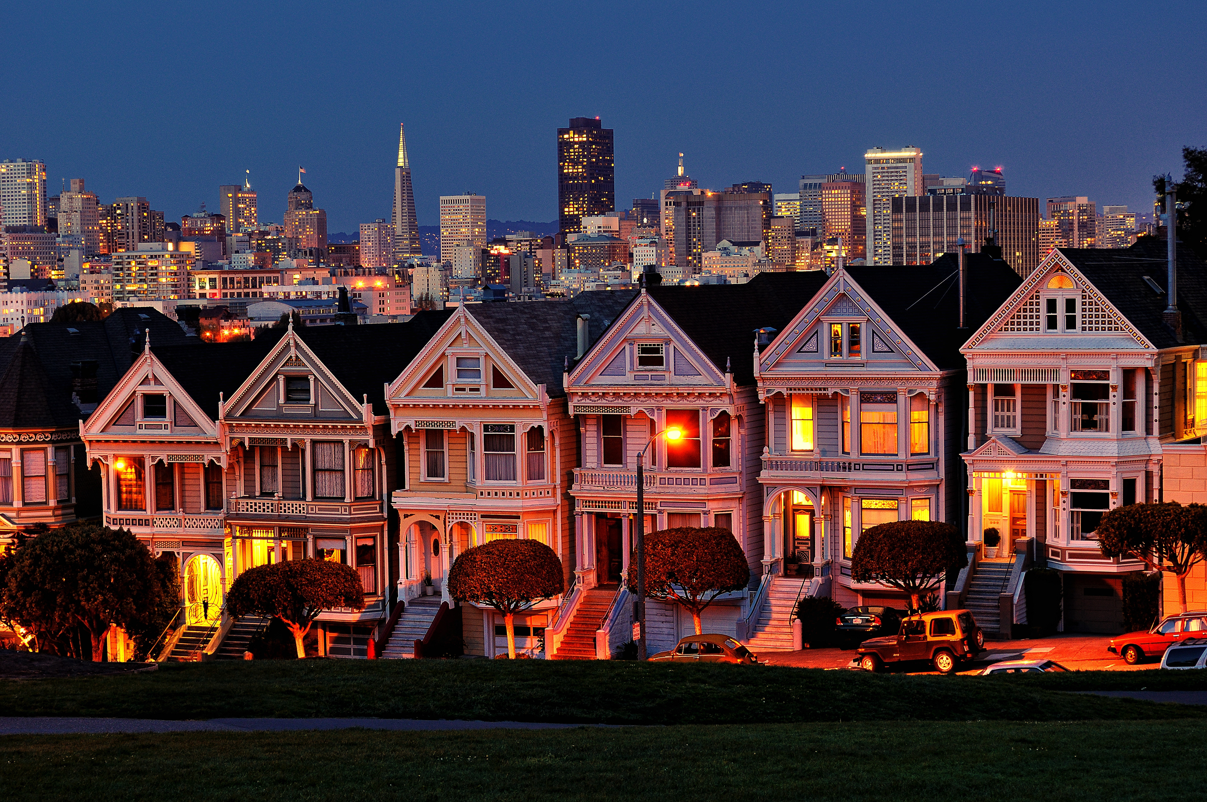 The iconic 'Painted Ladies' Victorian houses at Alamo Square, glowing warmly against the modern San Francisco skyline at dusk