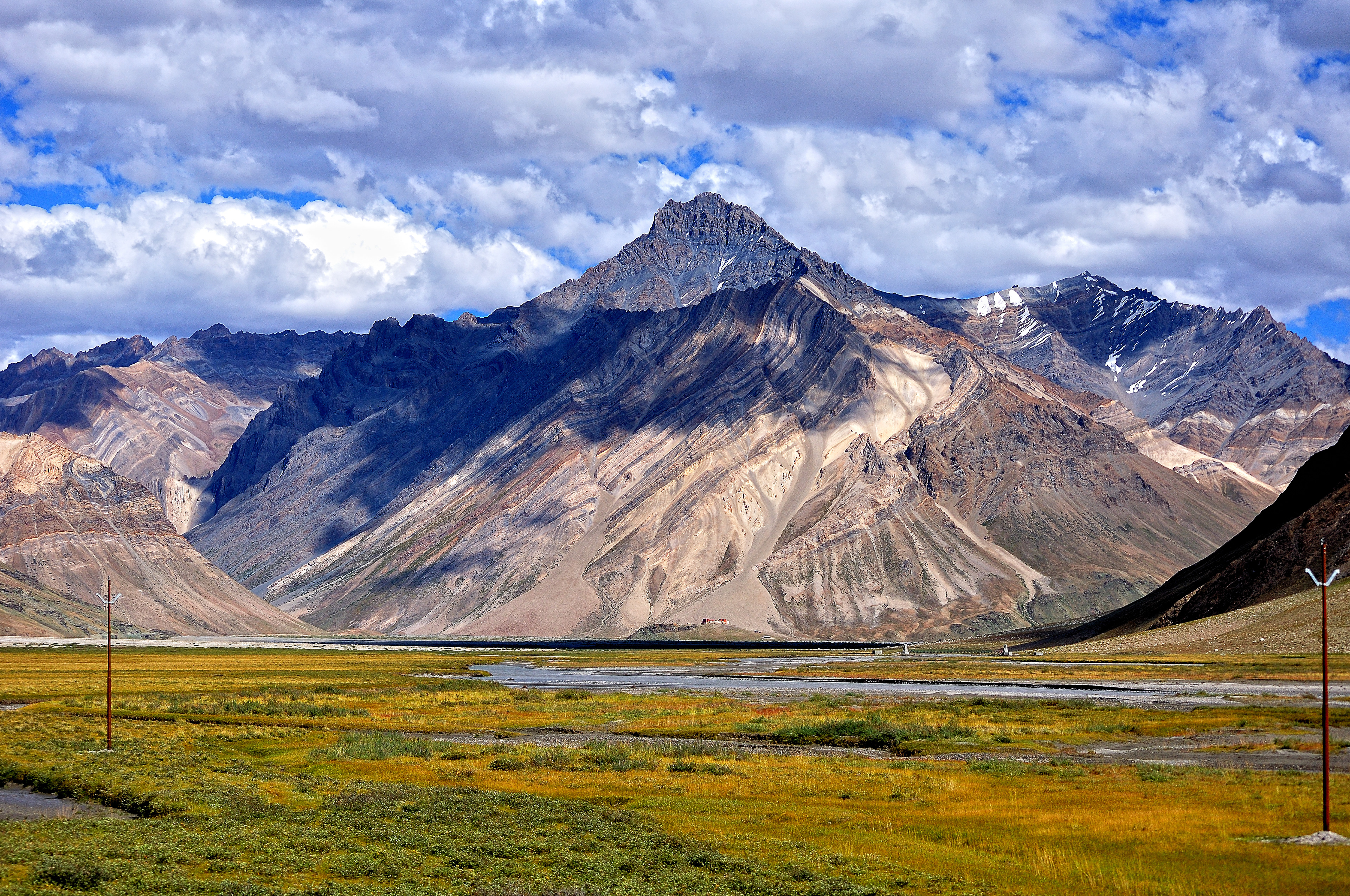 A vast green meadow in Rangdum, Suru Valley, framed by the towering, multi-colored rock formations of the Zanskar Range under a dramatic sky