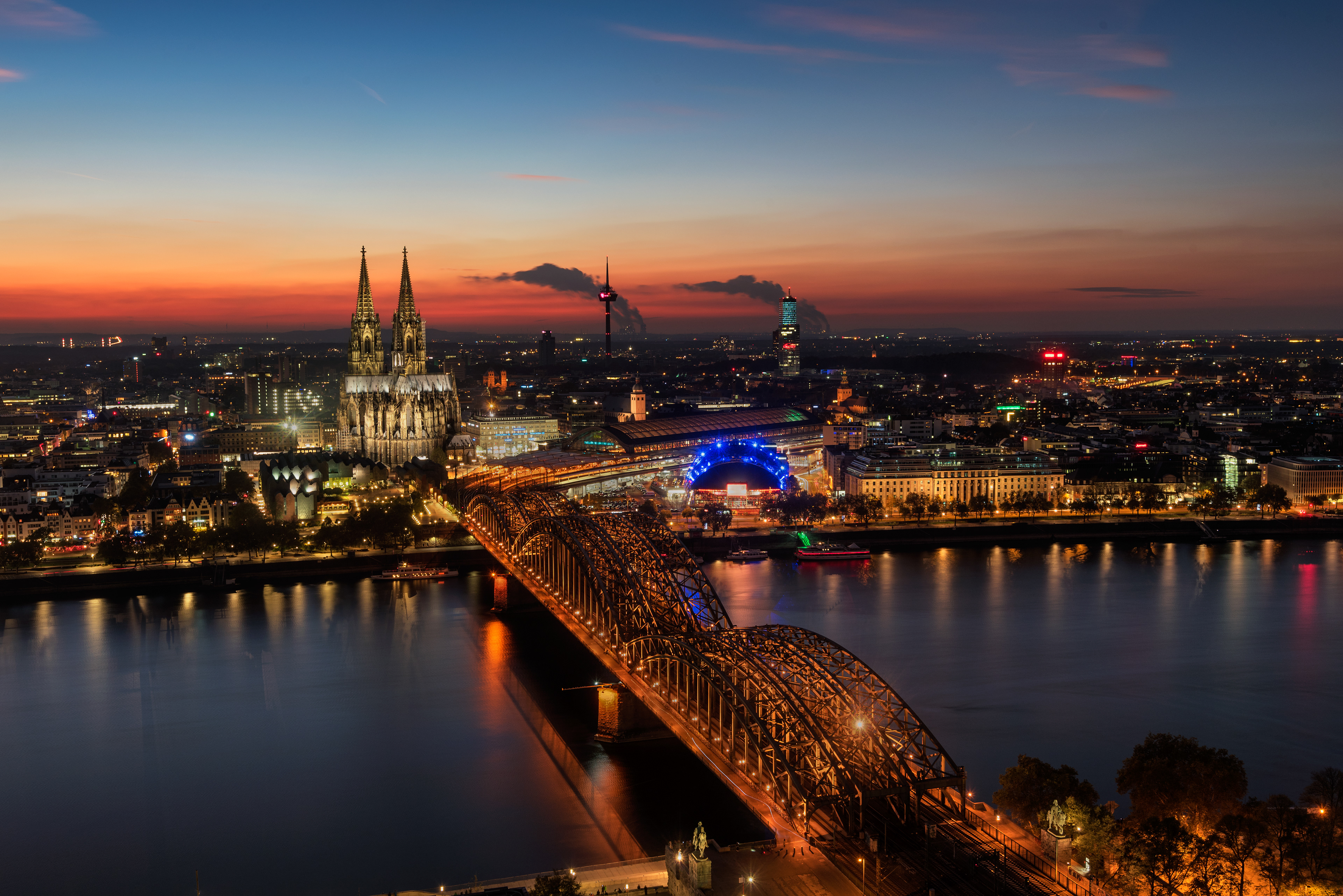 The twin spires of Cologne Cathedral and the Hohenzollern Bridge dominating the skyline above the Rhine River at sunset