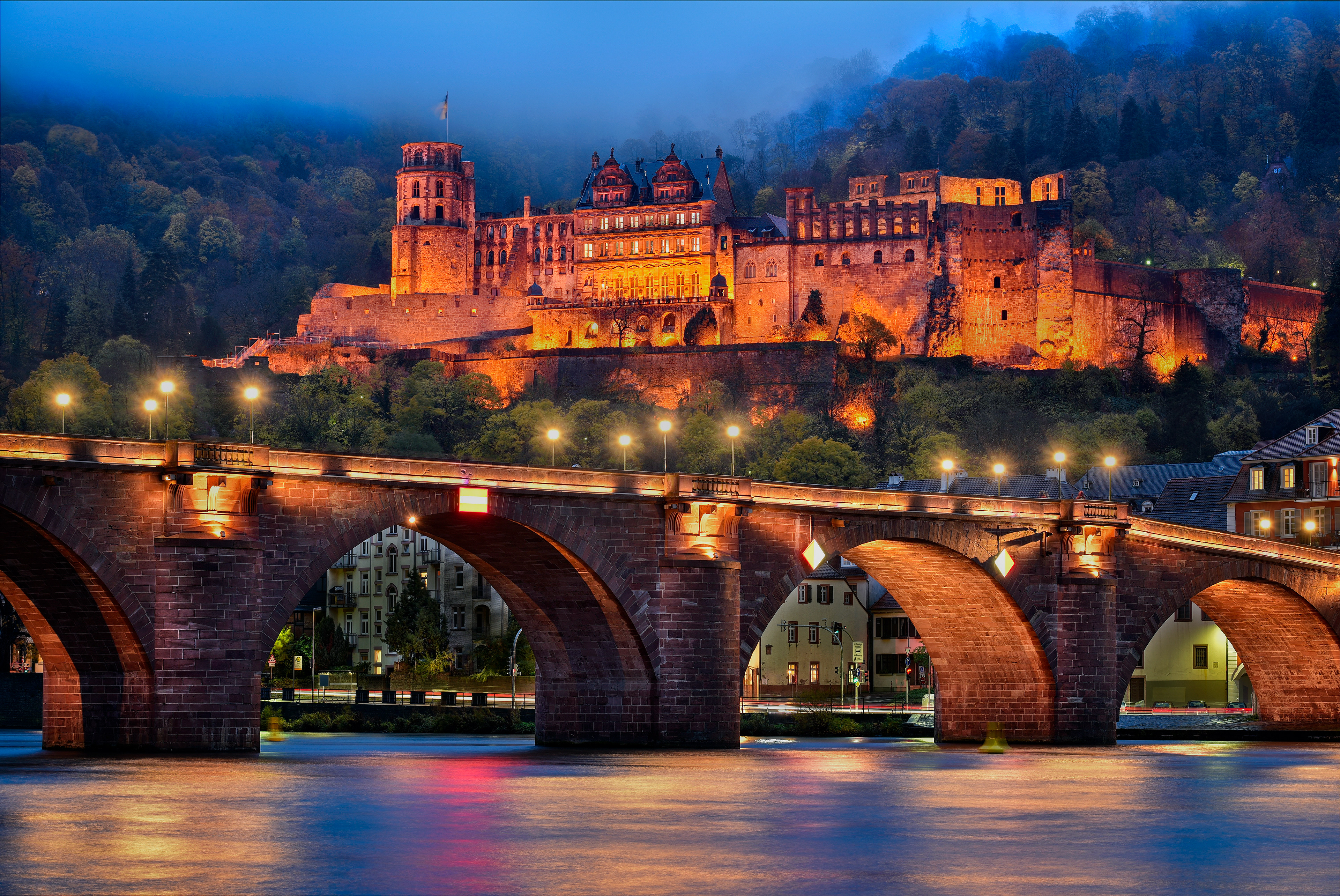 Heidelberg Castle glowing warmly on the hillside above the Neckar River, with the Old Bridge (Alte Brücke) illuminated in the foreground