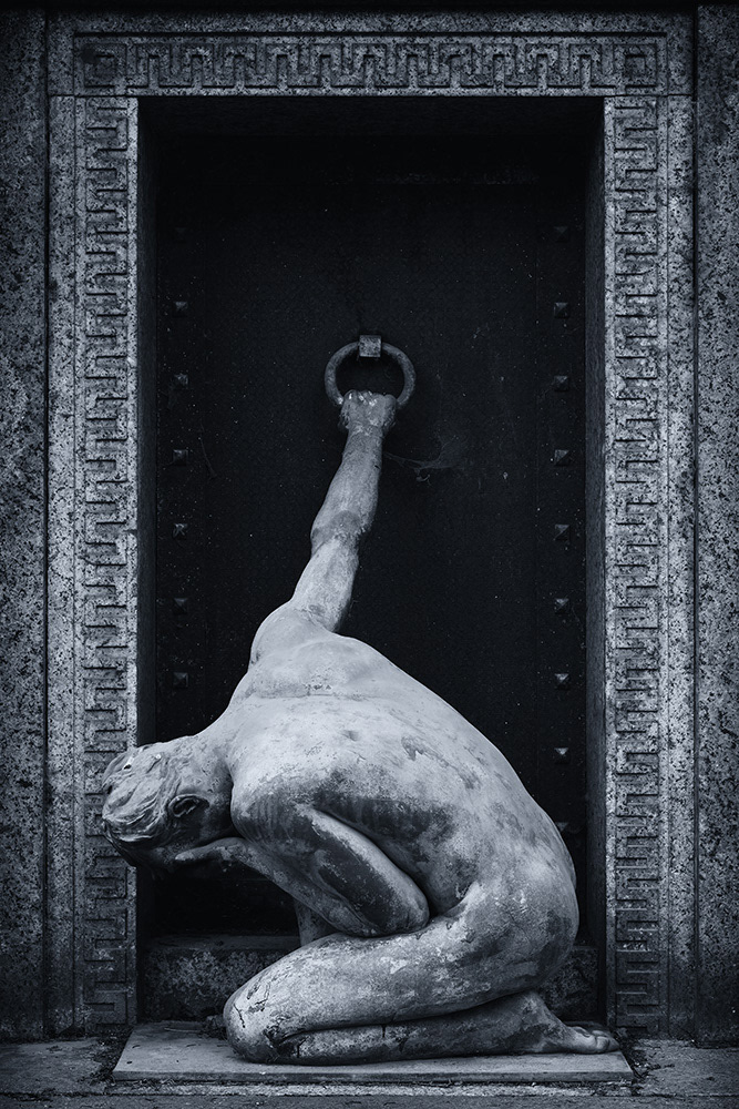 An unclothed athletic man kneels desperately in front of the door of a cemetery crypt, trying to gain access.