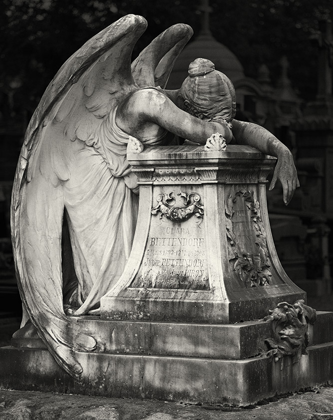 A female angel with bare shoulders and enormous wings has sadly lowered her head onto a monumental gravestone.