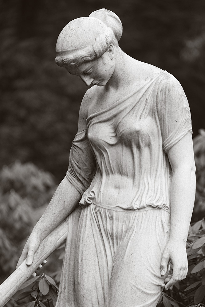 Sculpture of a young lady with a hair knot, bare shoulder and tight, transparent garment, standing in front of a grave.