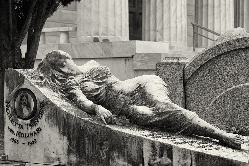 A sleeping young lady with bare shoulders, wearing a tight-fitting dress, holding a crucifix, in a cemetery in Genoa.