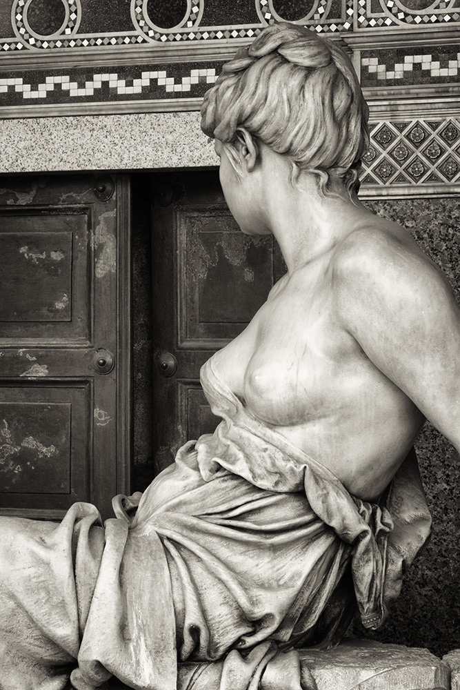 Erotic grave sculpture. Elegant lady with exposed breasts and gently flowing robe looks towards an open door of a crypt.