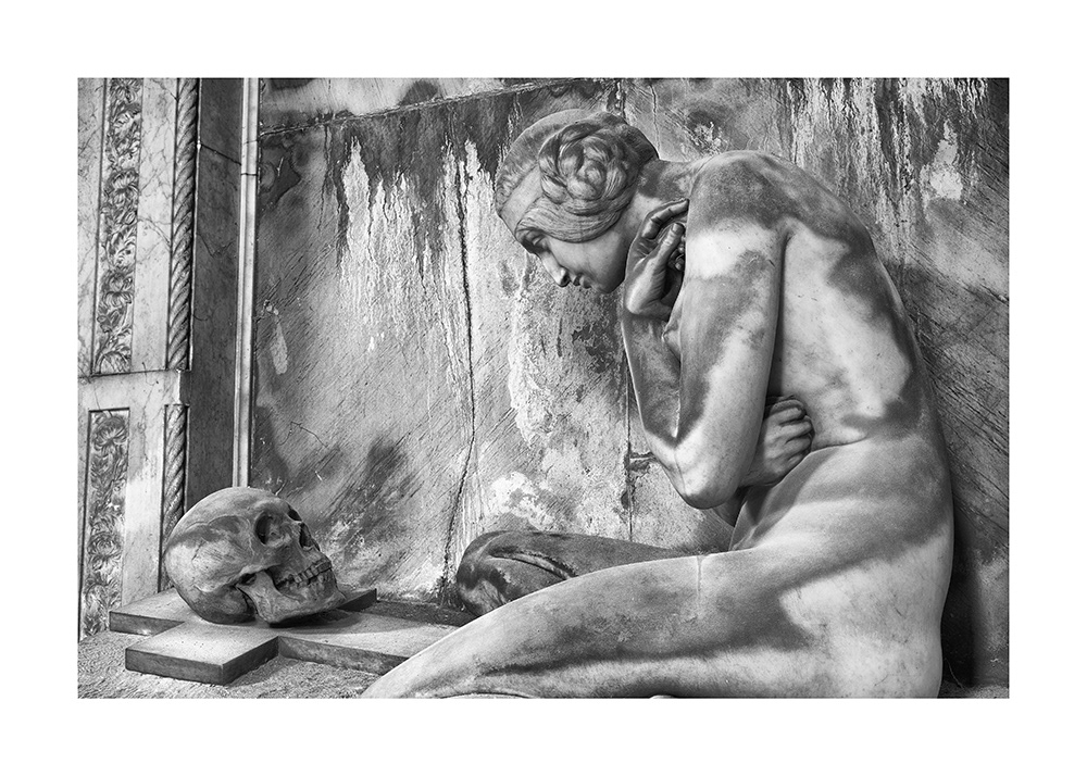 A naked woman kneels with a thoughtful face before a grave cross with a human skull in its center.
