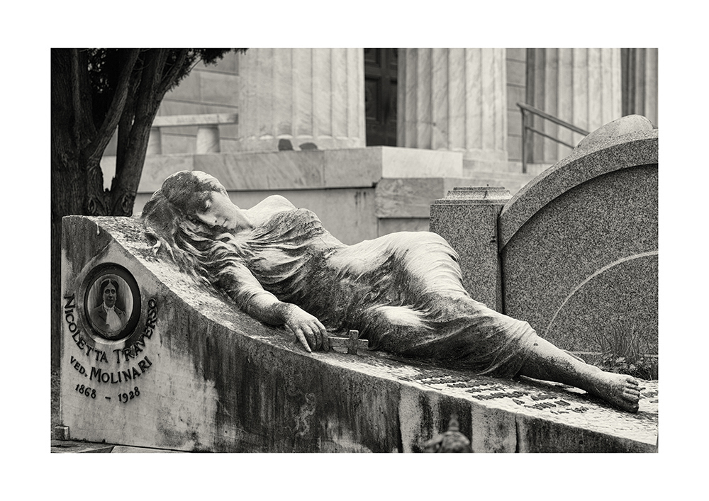 A sleeping young lady with bare shoulders, wearing a tight-fitting dress, holding a crucifix, in a cemetery in Genoa.