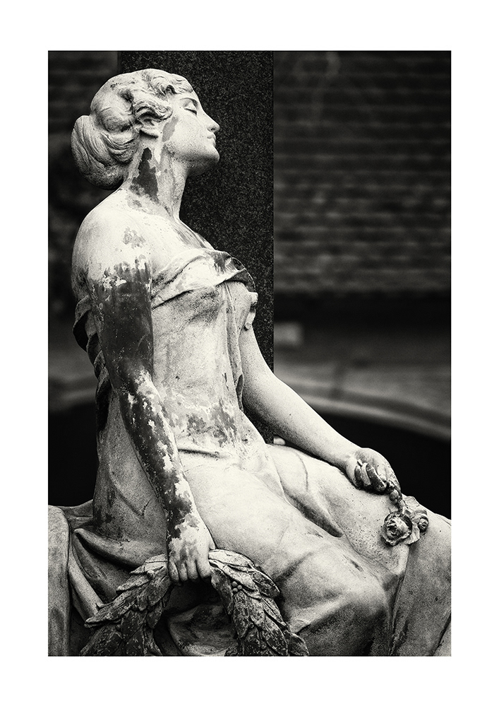 A graceful young lady sits with closed eyes and bare shoulders at a grave, holding roses and a funeral wreath in her hands.