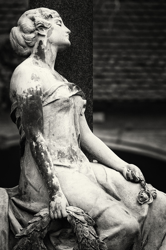 A graceful young lady sits with closed eyes and bare shoulders at a grave, holding roses and a funeral wreath in her hands.