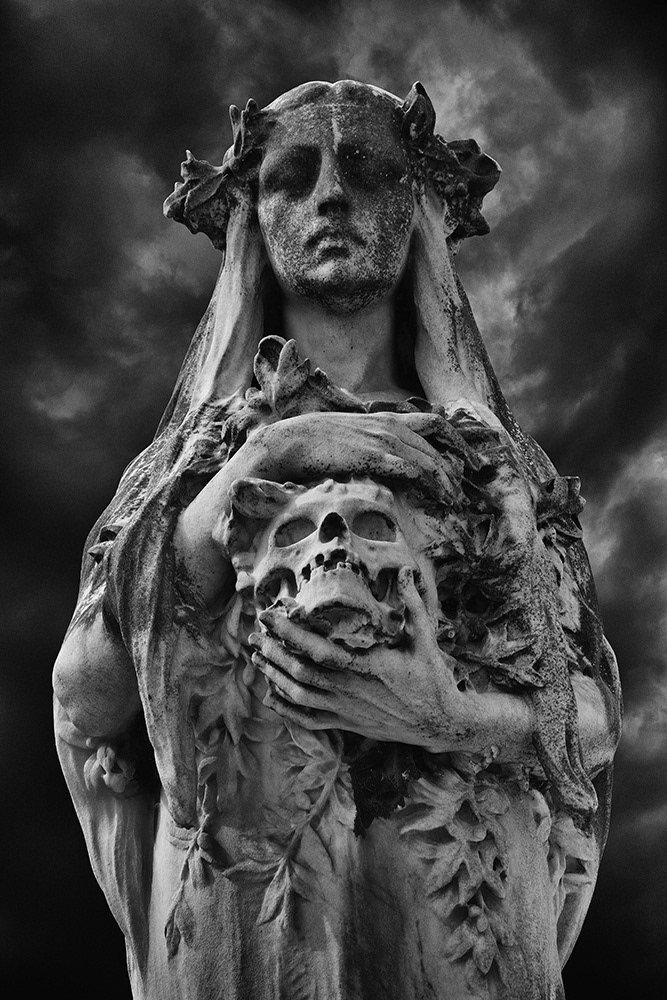 A female figure holds a florally decorated skull in her hands. Viewed from below against a dark stormy sky.