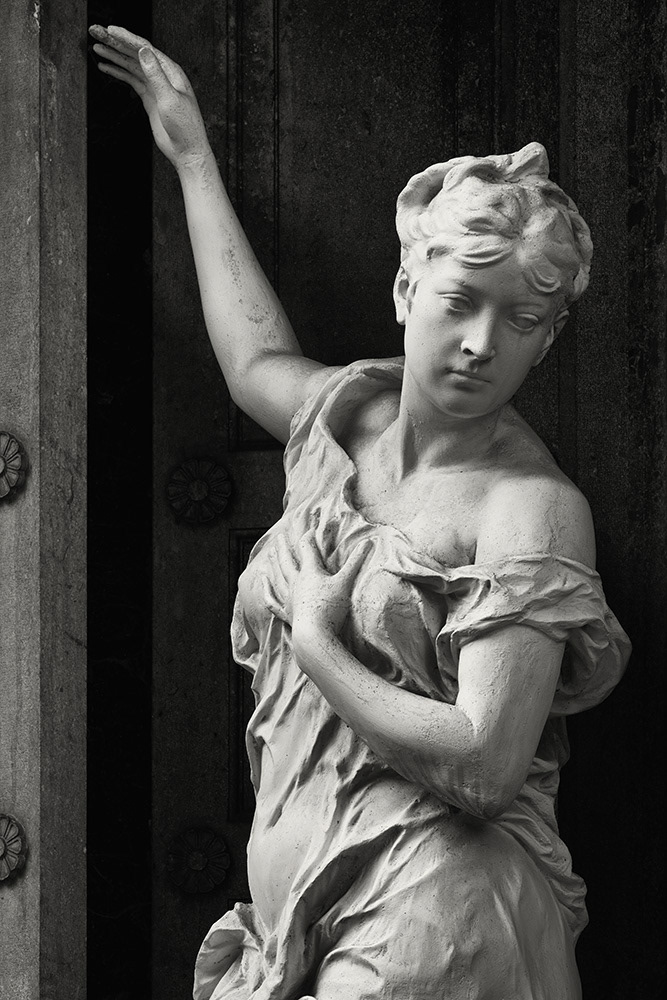 Sculpture of a female mourning figure with a shoulder-free dress in front of the false door of a crypt, inviting gesture.
