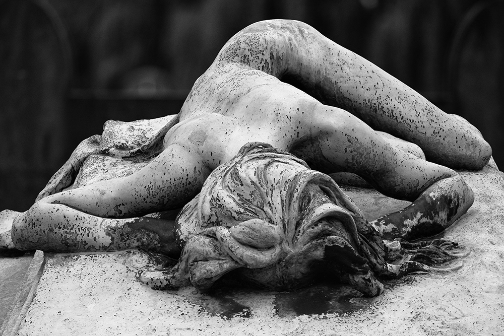 Cemetery sculpture of an unclothed woman with long hair lying on an inclined grave slab.