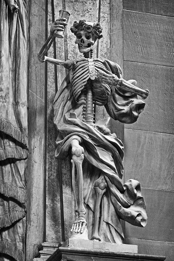 Death, a skeleton in robes, wearing a headdress of roses and ears of grain, holds an hourglass aloft in a warning gesture.