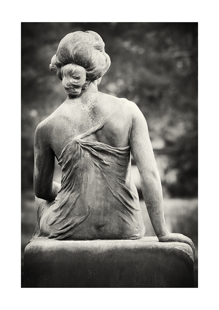 Rear view of a slim young woman with bare shoulders and a transparent dress, sitting on a gravestone.
