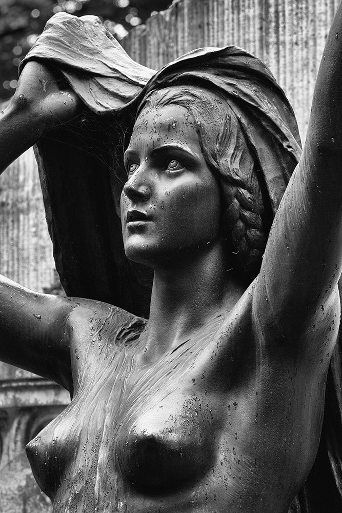Sculpture of a naked young lady in the rain, holding a cloth protectively over her head, made of black patinated bronze.