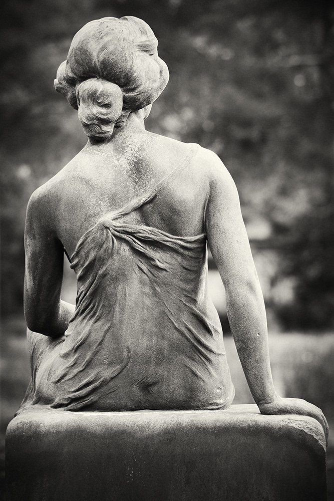 Rear view of a slim young woman with bare shoulders and a transparent dress, sitting on a gravestone.