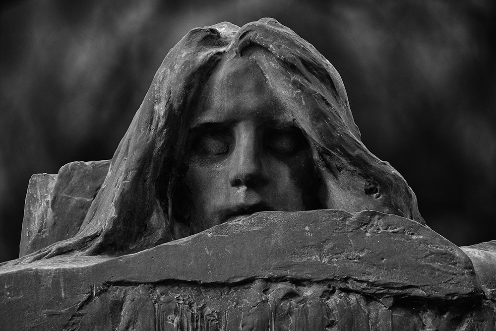 The grim face of death with long hair and deep-set, closed eyes appears lurking behind a tombstone.