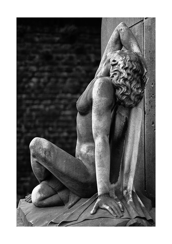 Sculpture of a slim, unclothed young woman with long, curly hair, sitting on a gravesite. The gaze is directed skywards.