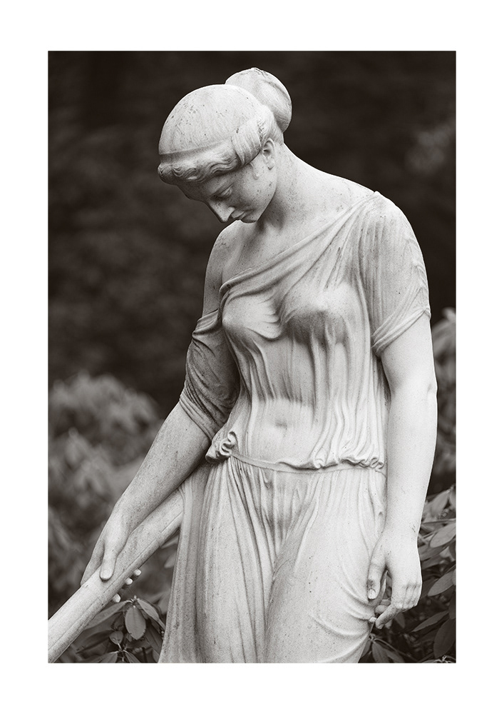 Sculpture of a young lady with a hair knot, bare shoulder and tight, transparent garment, standing in front of a grave.