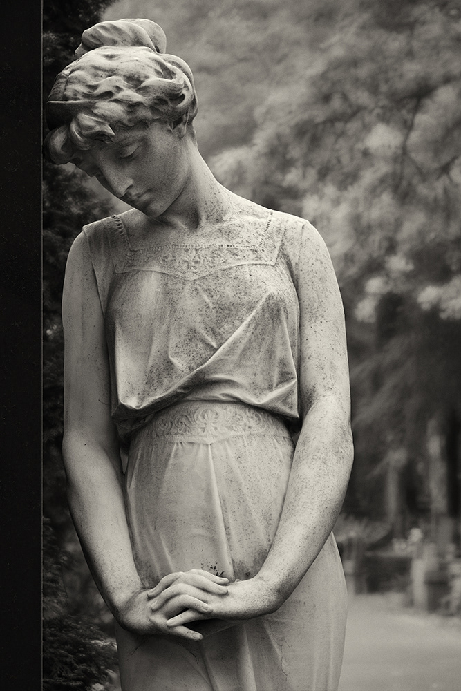 Sculpture of a young, graceful woman in a lace dress, her head bowed and resting against a tombstone, her hands intertwined.