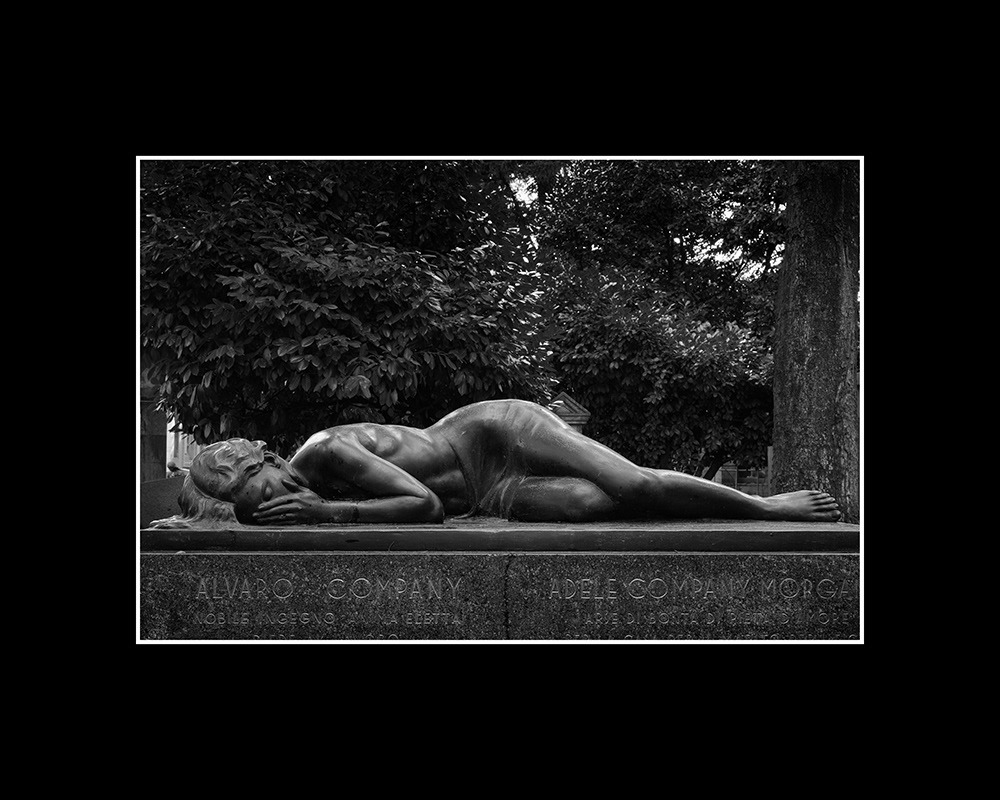 A sensual female body, discreetly draped only around the lower abdomen, lies asleep on a tombstone in Milan.