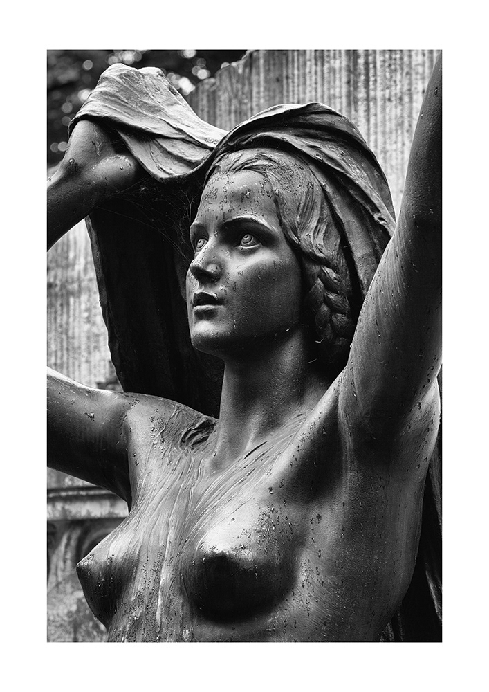 Sculpture of a naked young lady in the rain, holding a cloth protectively over her head, made of black patinated bronze.