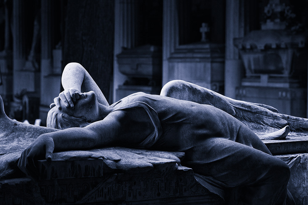 A dreaming female angel lies on a gravestone in Genoa, with a colonnade containing more graves in the background.