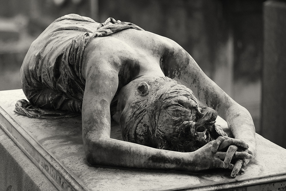 A slender young lady lies with her shoulders bare on the lid of a sarcophagus in a Parisian cemetery.