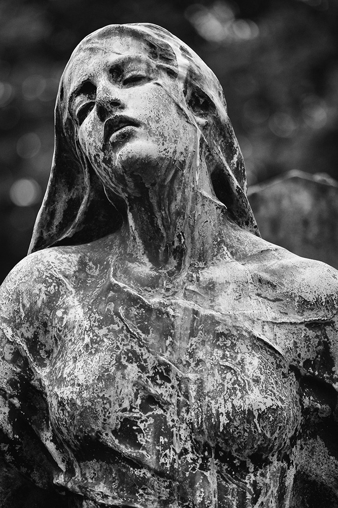 Bronze sculpture of a young lady in a Parisian cemetery, half-closed eyes, slightly open mouth, covered in patina.