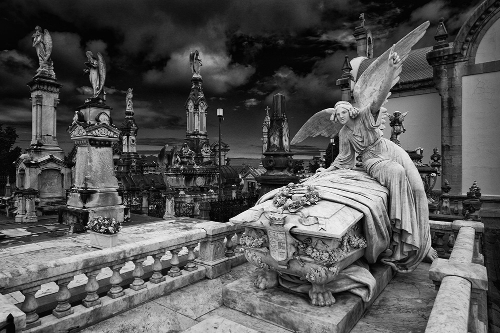 An angel guards a stone sarcophagus; next to it, various tombs rise steeply against a dark sky.