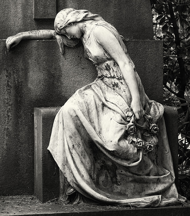 A beautiful young woman with bare shoulders, a long dress and a headscarf leans against a gravestone in deep sorrow.