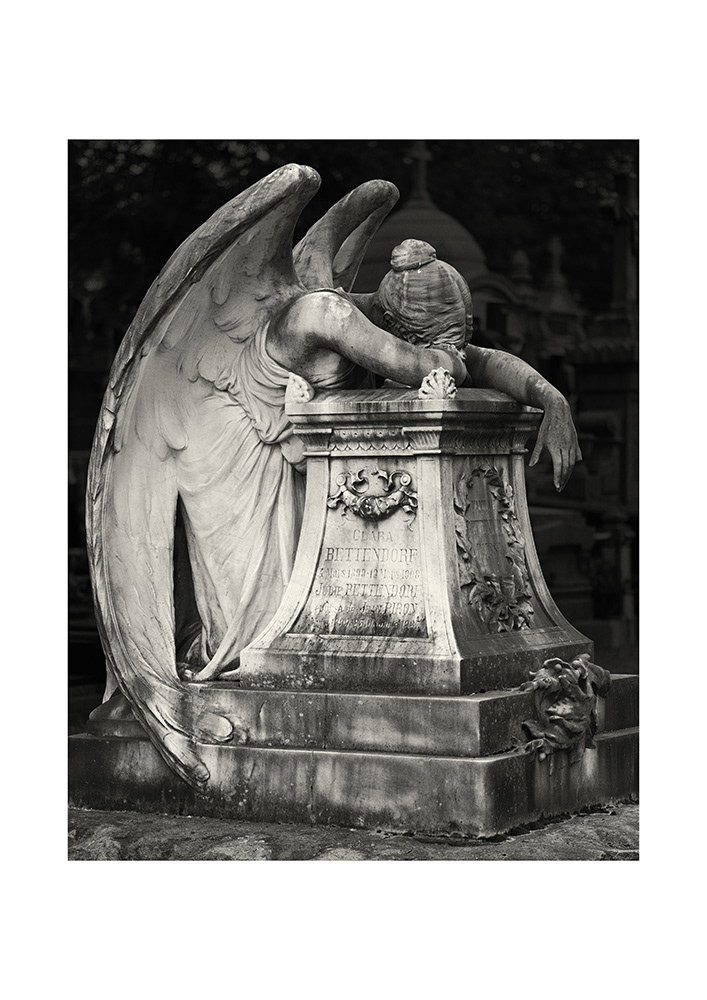 A female angel with bare shoulders and enormous wings has sadly lowered her head onto a monumental gravestone.