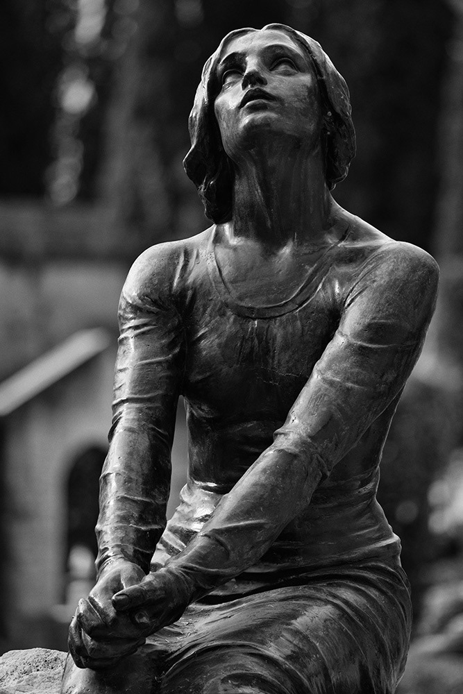 A young woman sits on a gravestone with her hands clasped together, her gaze directed expectantly towards the sky.