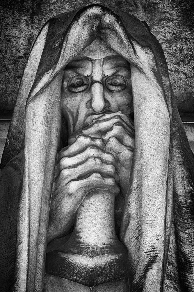 Man with piercing eyes, frowning face and bony hands, head covered by a hood, leaning on his sword.