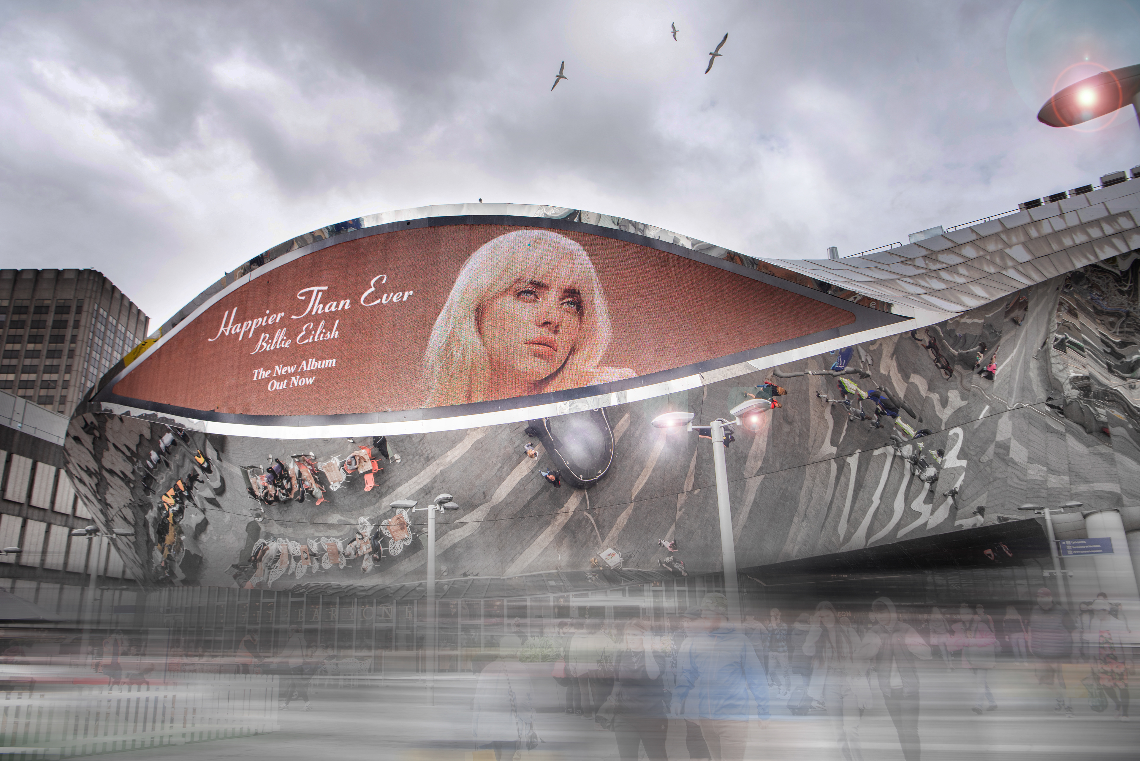 Modern building with a large billboard featuring "Happier Than Ever" album, an image of a woman, and reflected pedestrians below. Birds soar in a cloudy sky.