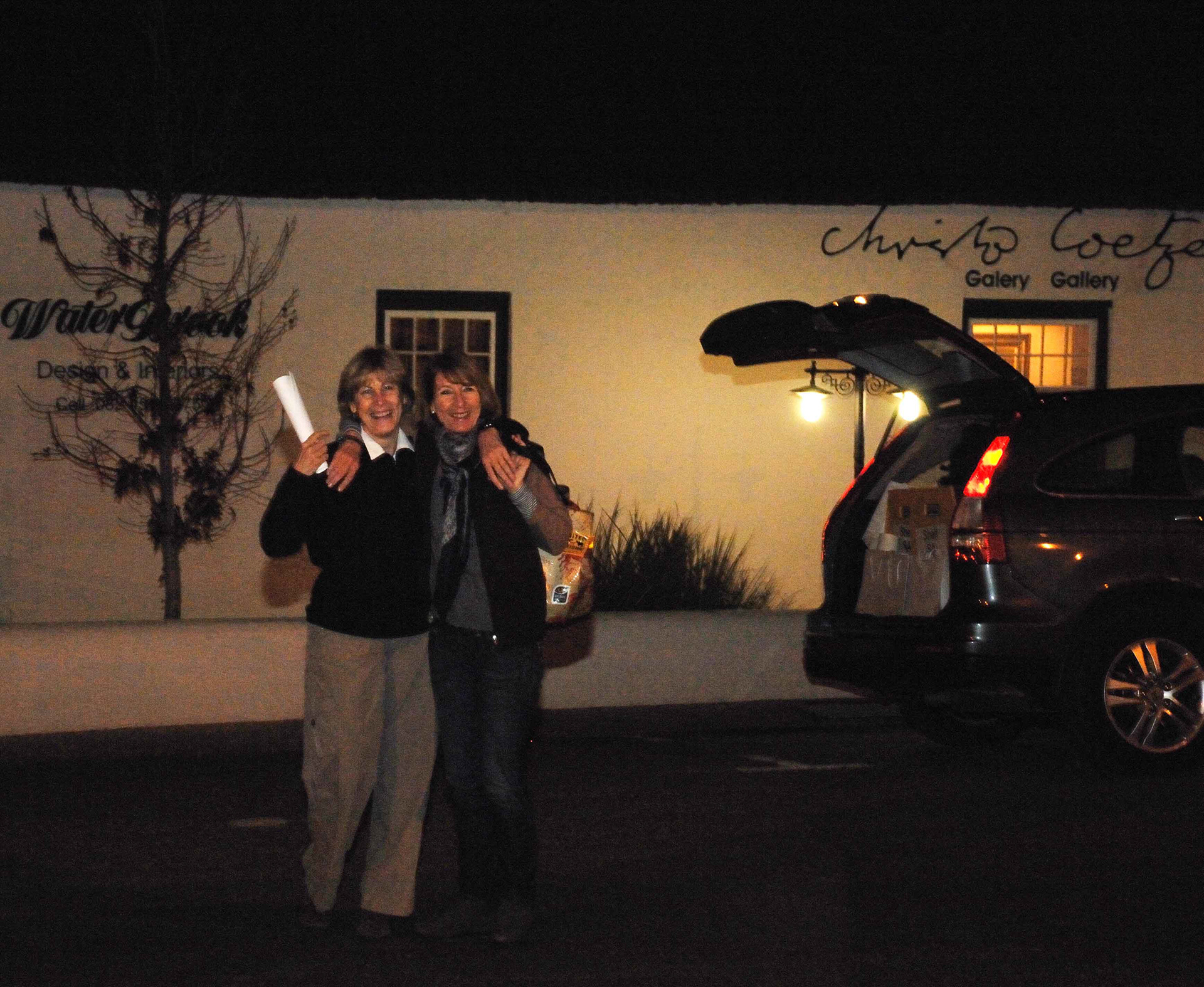 Two people happily embrace beside a car with an open trunk at night. They stand outside under a warm light, conveying a sense of joy and friendship.