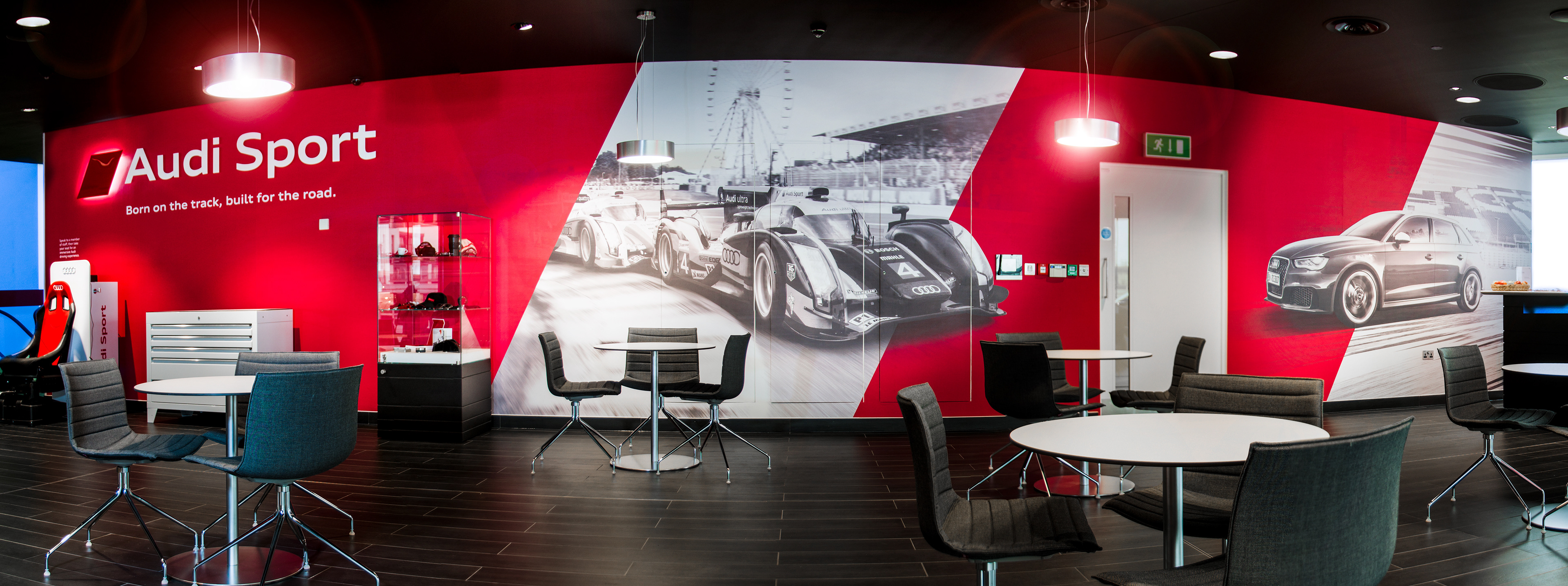 Modern lounge with red walls featuring large images of Audi race and sport cars. "Audi Sport" logo displayed. Sleek tables and chairs create a dynamic, racing-inspired atmosphere.