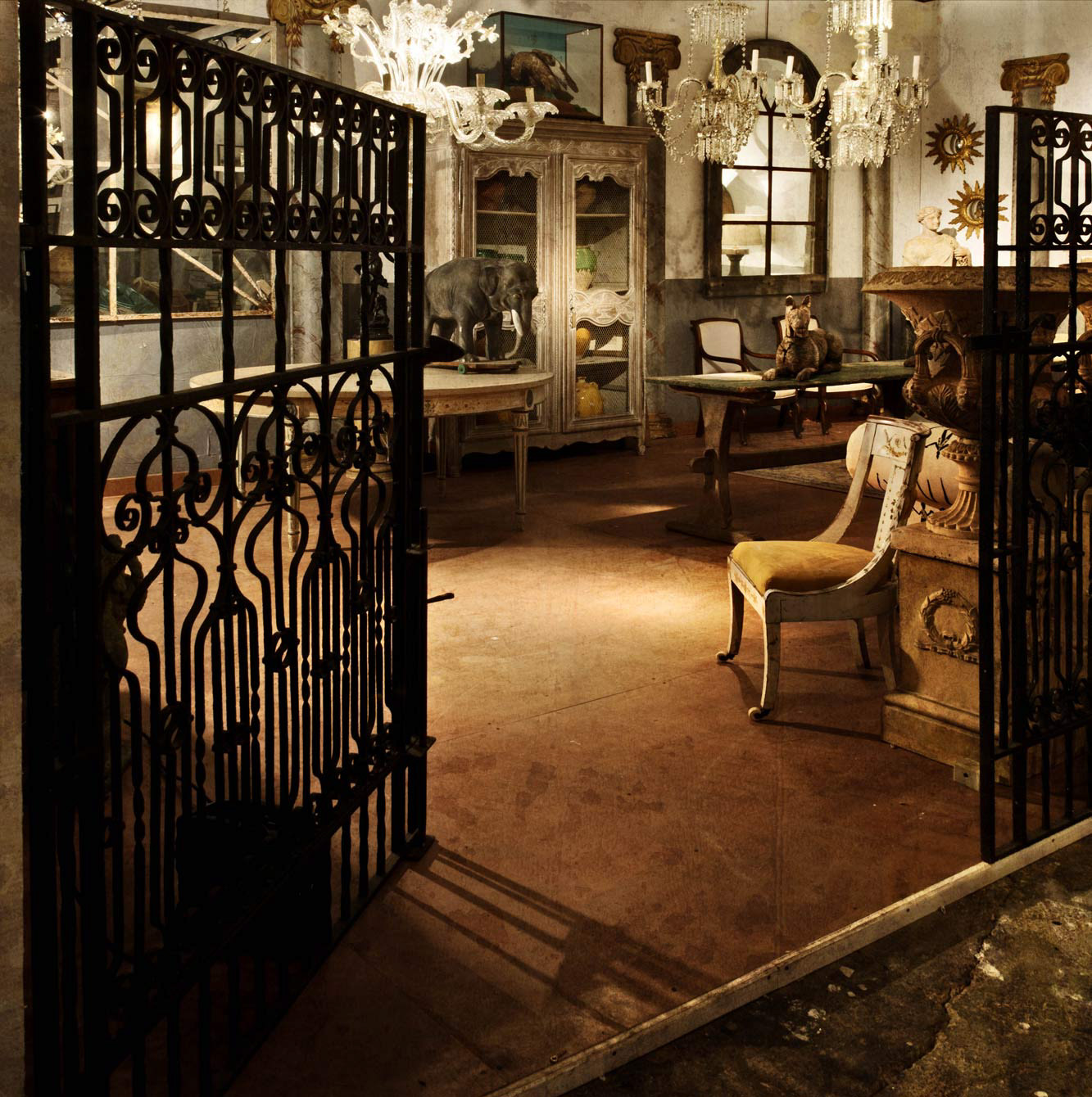 Dimly lit vintage room with ornate iron gates, chandeliers, wooden furniture, and animal sculptures. The setting feels rustic and elegant.