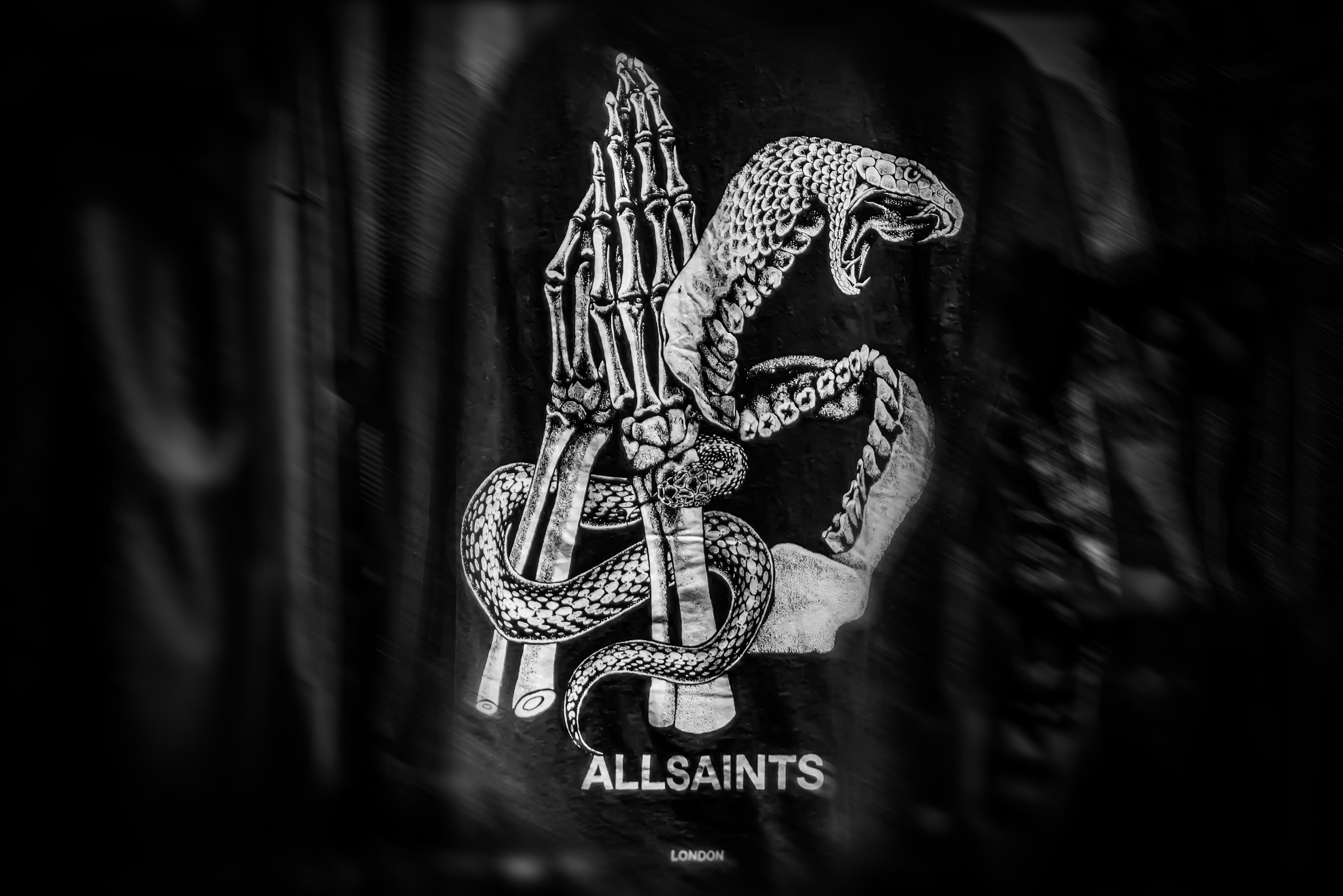 A skeletal hand and a coiled snake intertwine in a monochrome design on a dark background. Below, the text reads "AllSaints London," conveying a gothic tone.