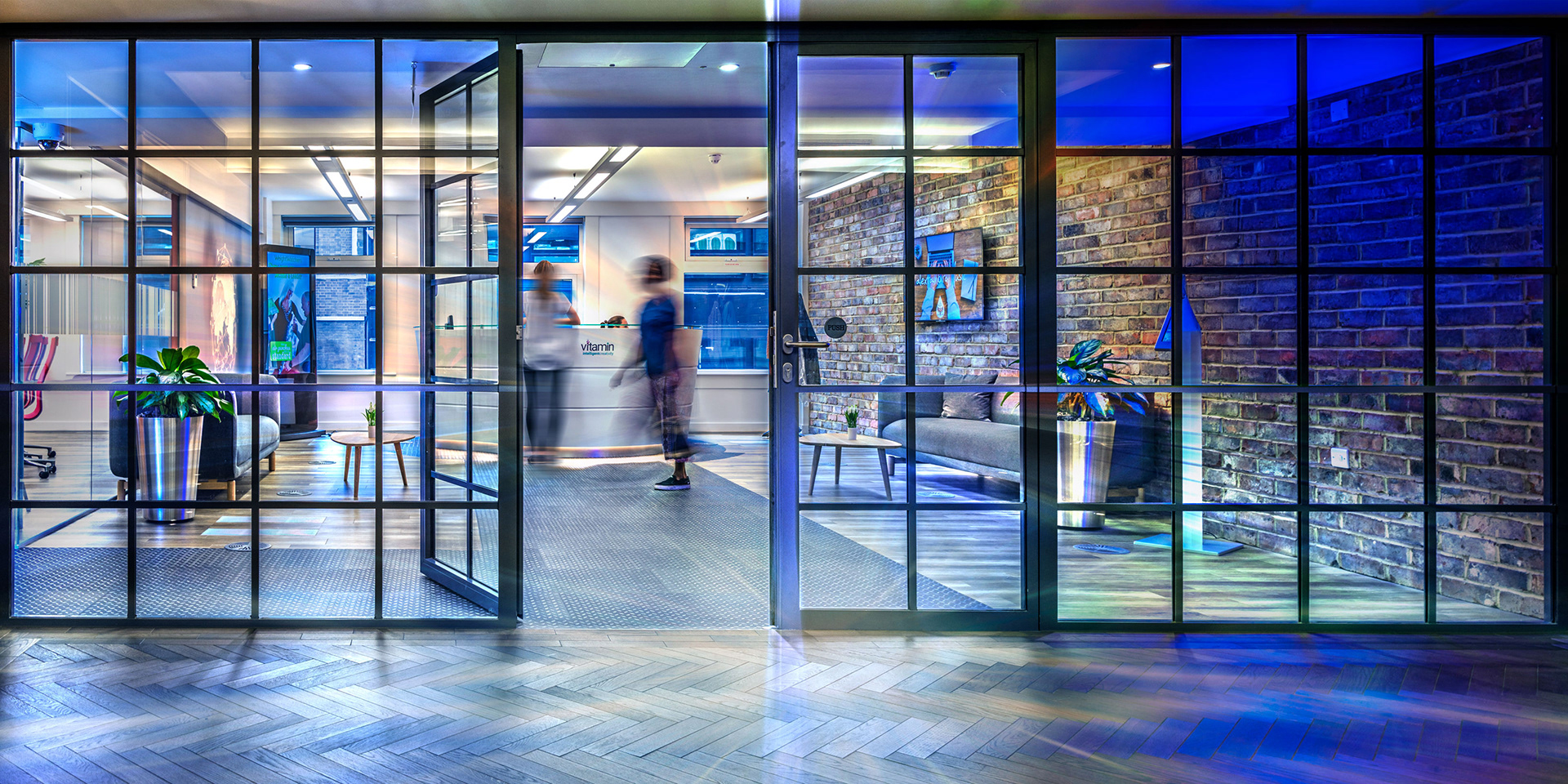 Modern office lobby with glass doors, exposed brick walls, and wooden floors. People walk by reception, creating a dynamic, contemporary atmosphere.