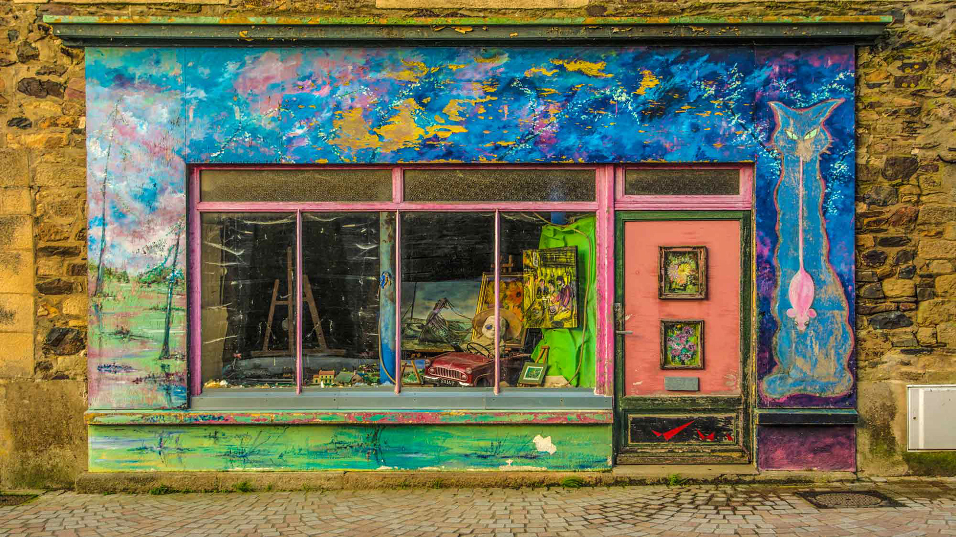 Colorful storefront with vibrant blue and pink mural, featuring a cat silhouette. Artworks are displayed in the window, set on cobblestone street.