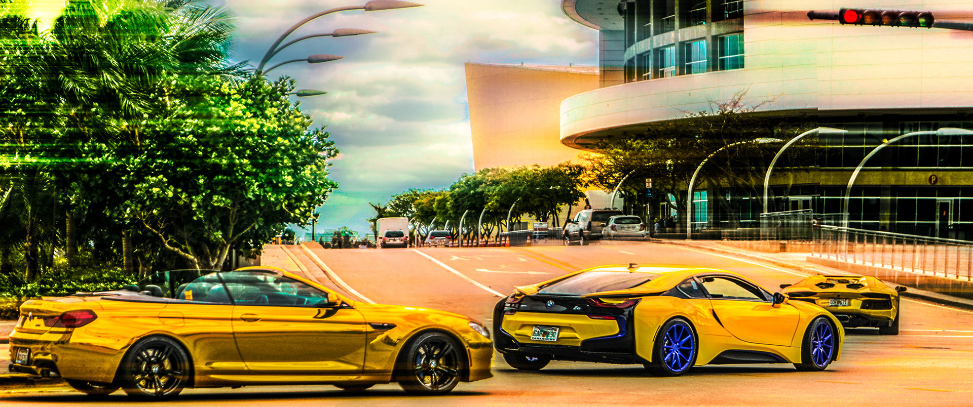 Vibrant scene of three shiny, gold sports cars with blue accents driving on a city street, surrounded by lush green trees and modern buildings.