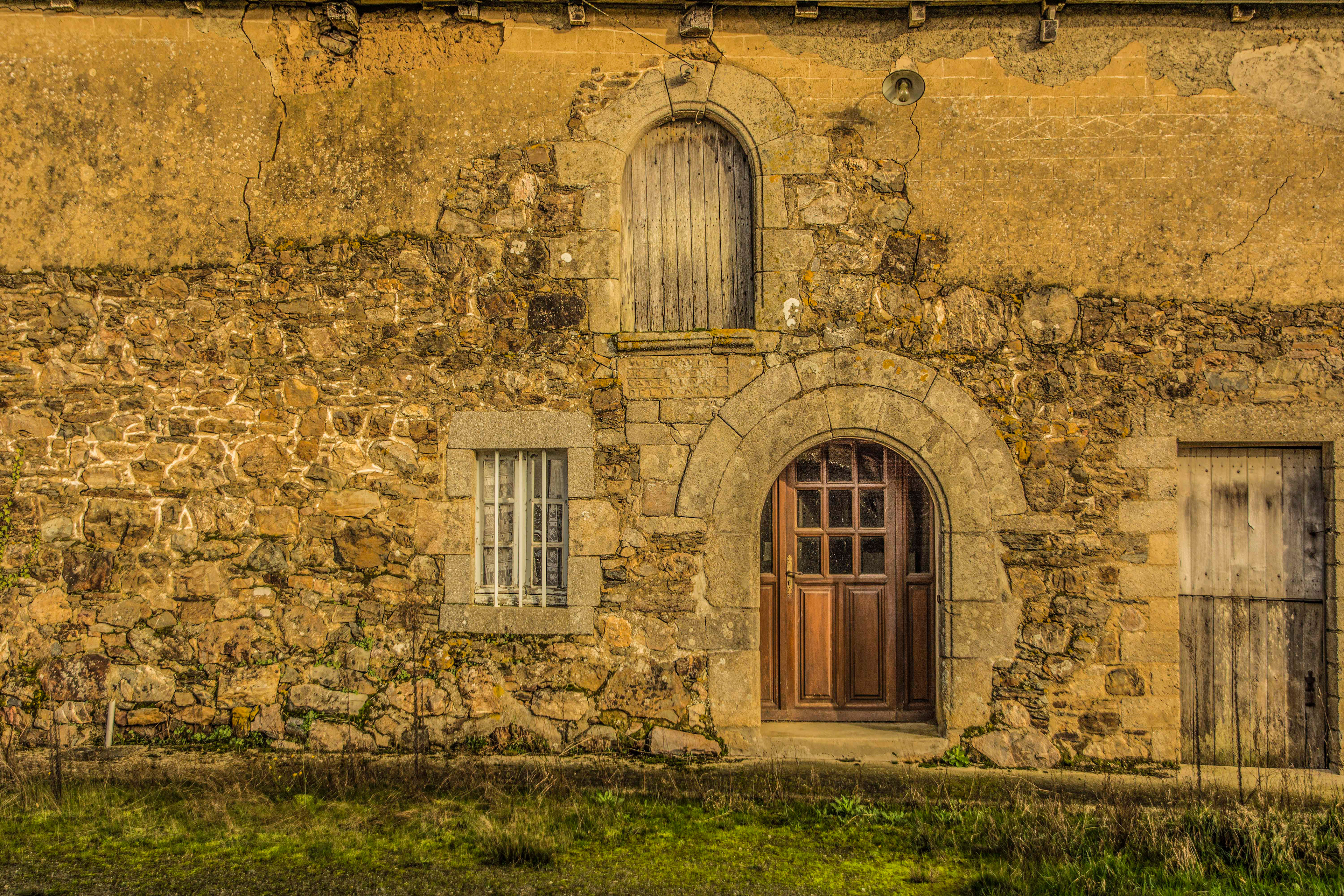 A rustic stone building with a weathered wooden door and arched frame. A small window with bars and an overgrown grass foreground convey an aged charm.