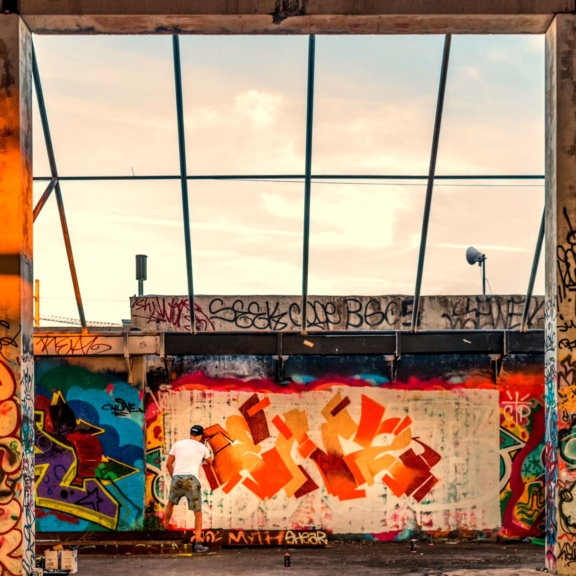 Graffiti artist spray painting vibrant orange abstract shapes on a wall covered in various tags. A warm, urban, and creative atmosphere.