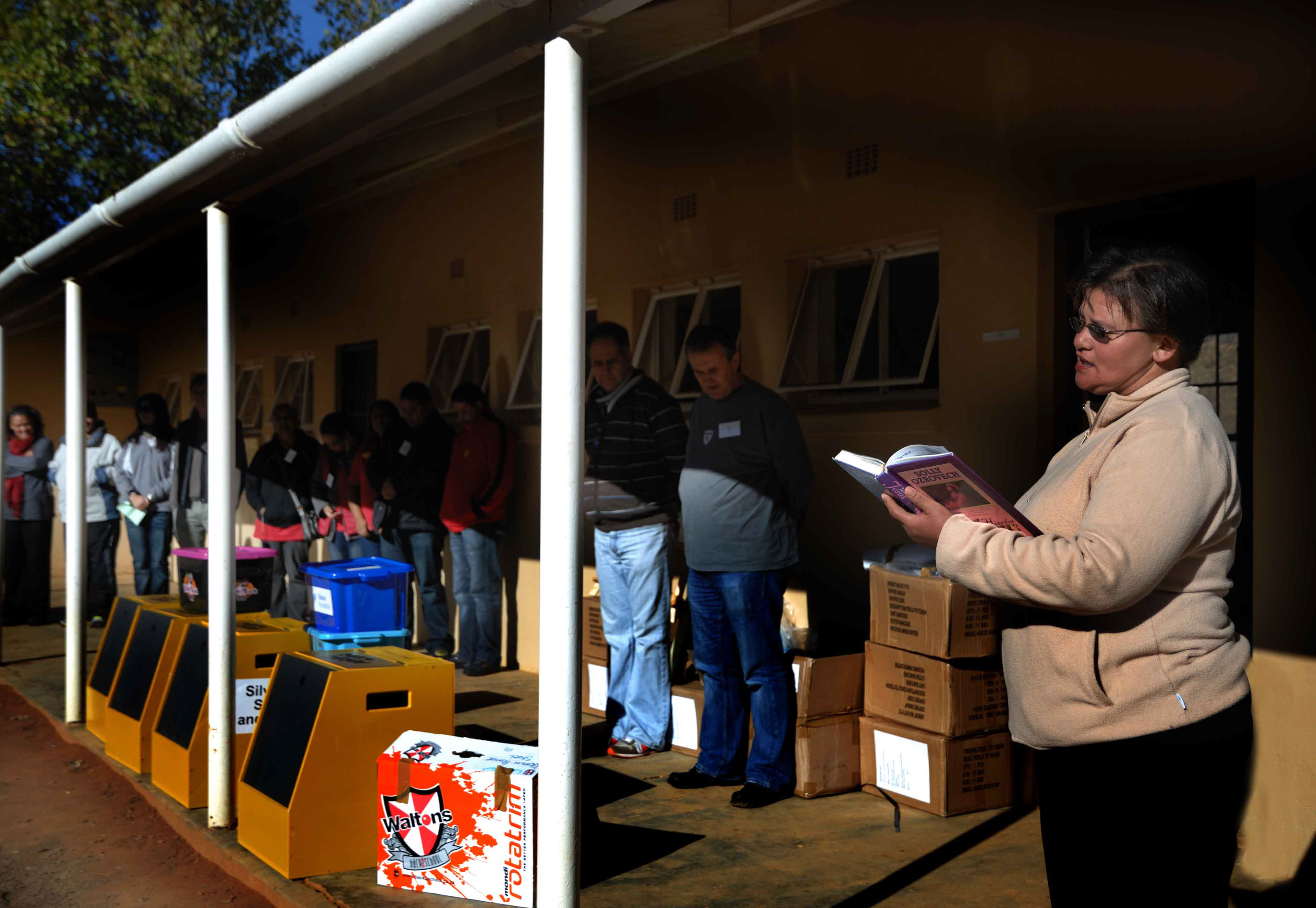 A group of people stands solemnly in a shaded area, facing yellow donation bins labeled "Silver Spec Savers." A woman in a beige jacket reads from a purple book, surrounded by cardboard boxes, conveying a sense of community and reflection.