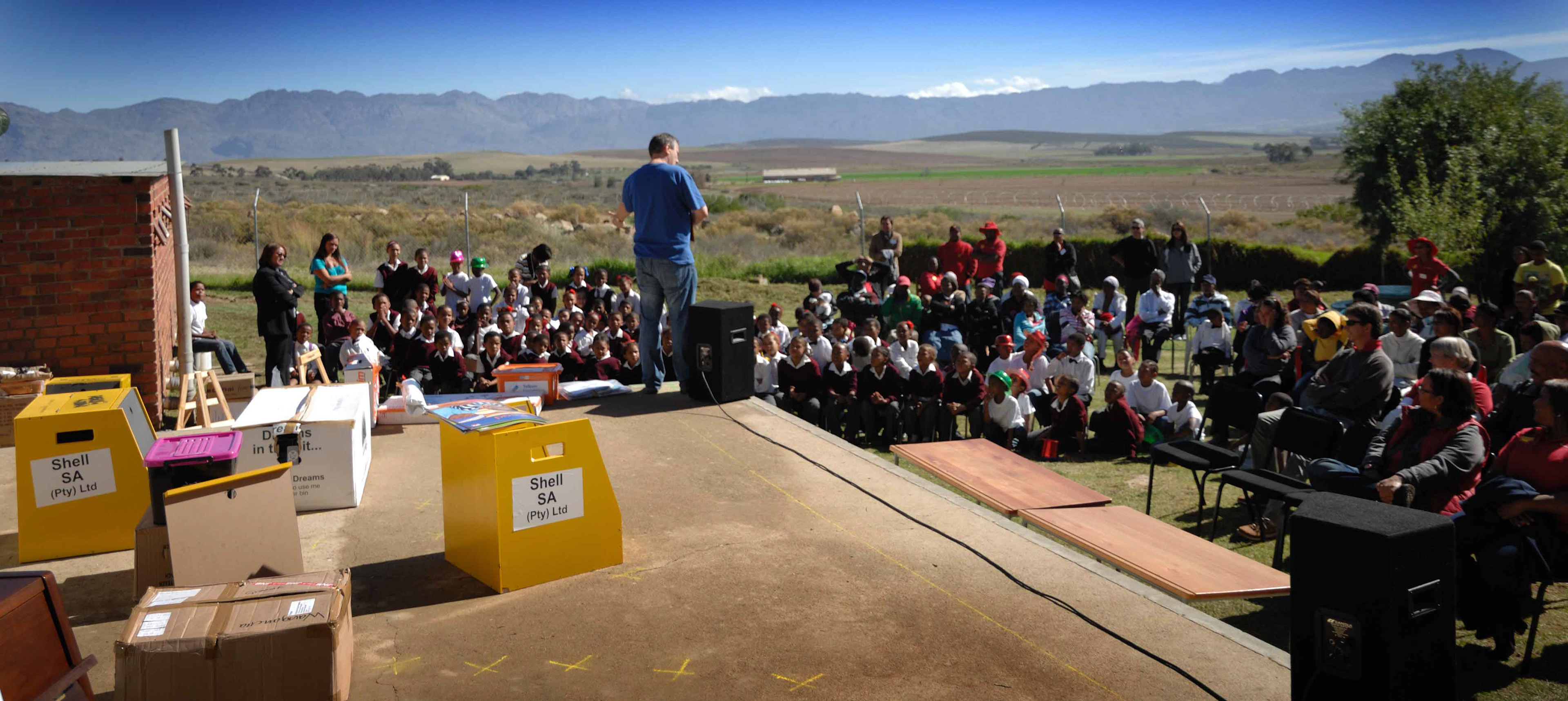 A man speaks to a large group of seated children and adults outdoors, with a scenic mountain view. Boxes labeled "Shell SA" are on a stage, conveying an educational event.