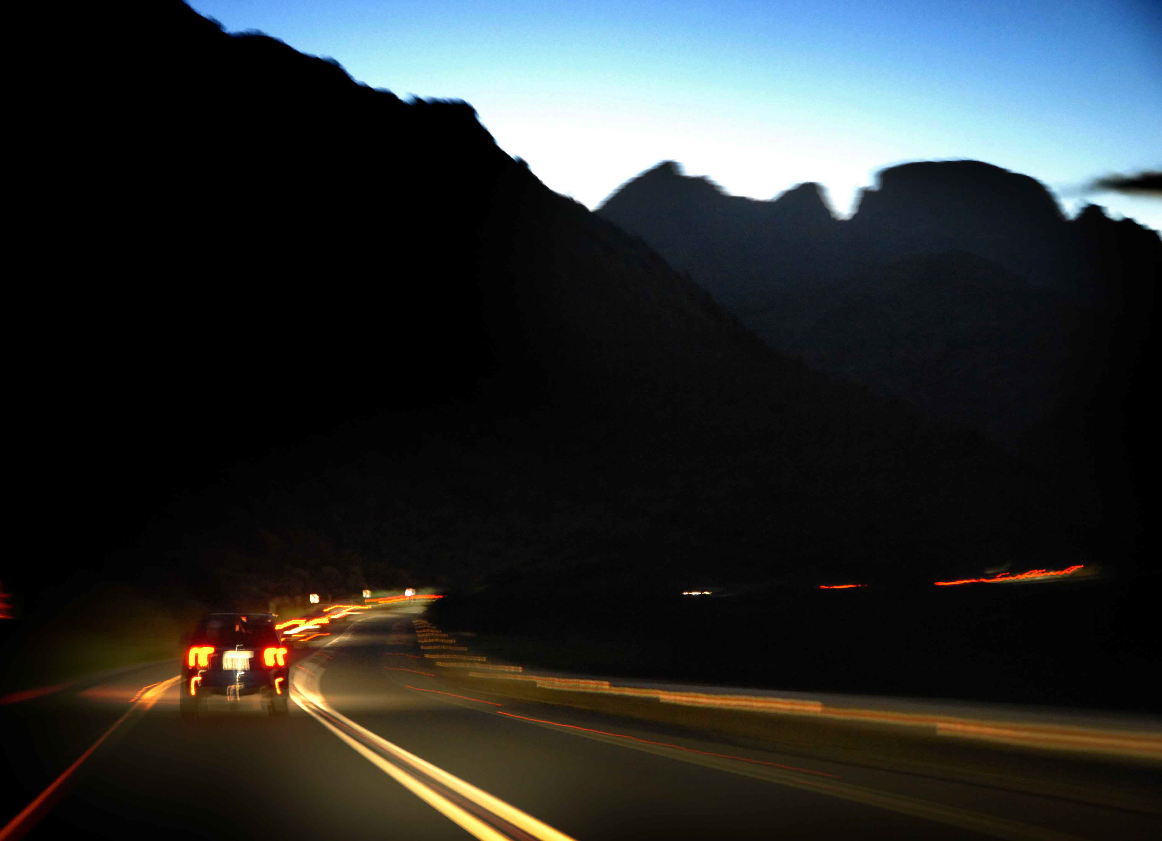 Blurred nighttime scene of a car driving on a winding road through dark mountains, with its taillights creating red streaks, evoking a sense of speed and solitude.