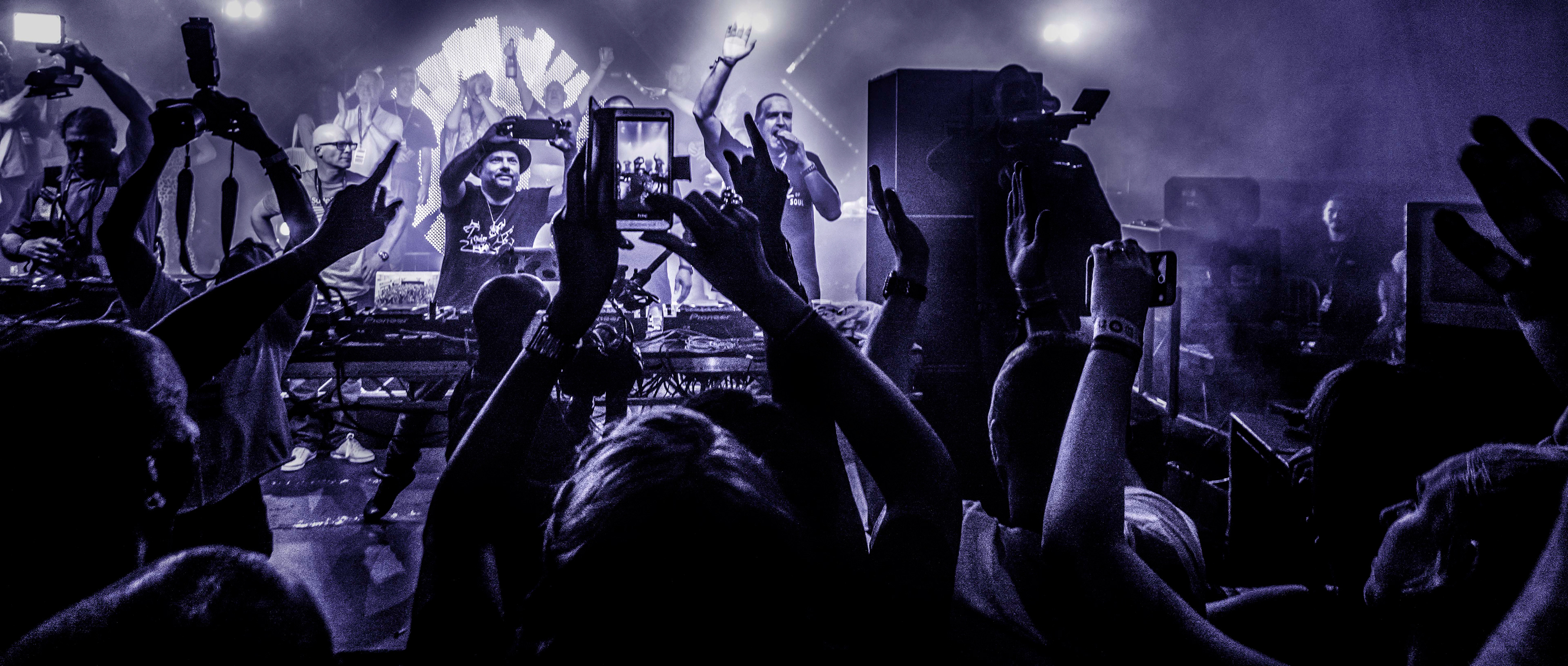 Concert scene with a DJ and rapper performing on stage, surrounded by a vibrant crowd under purple lighting. Enthusiastic fans raise phones and hands.