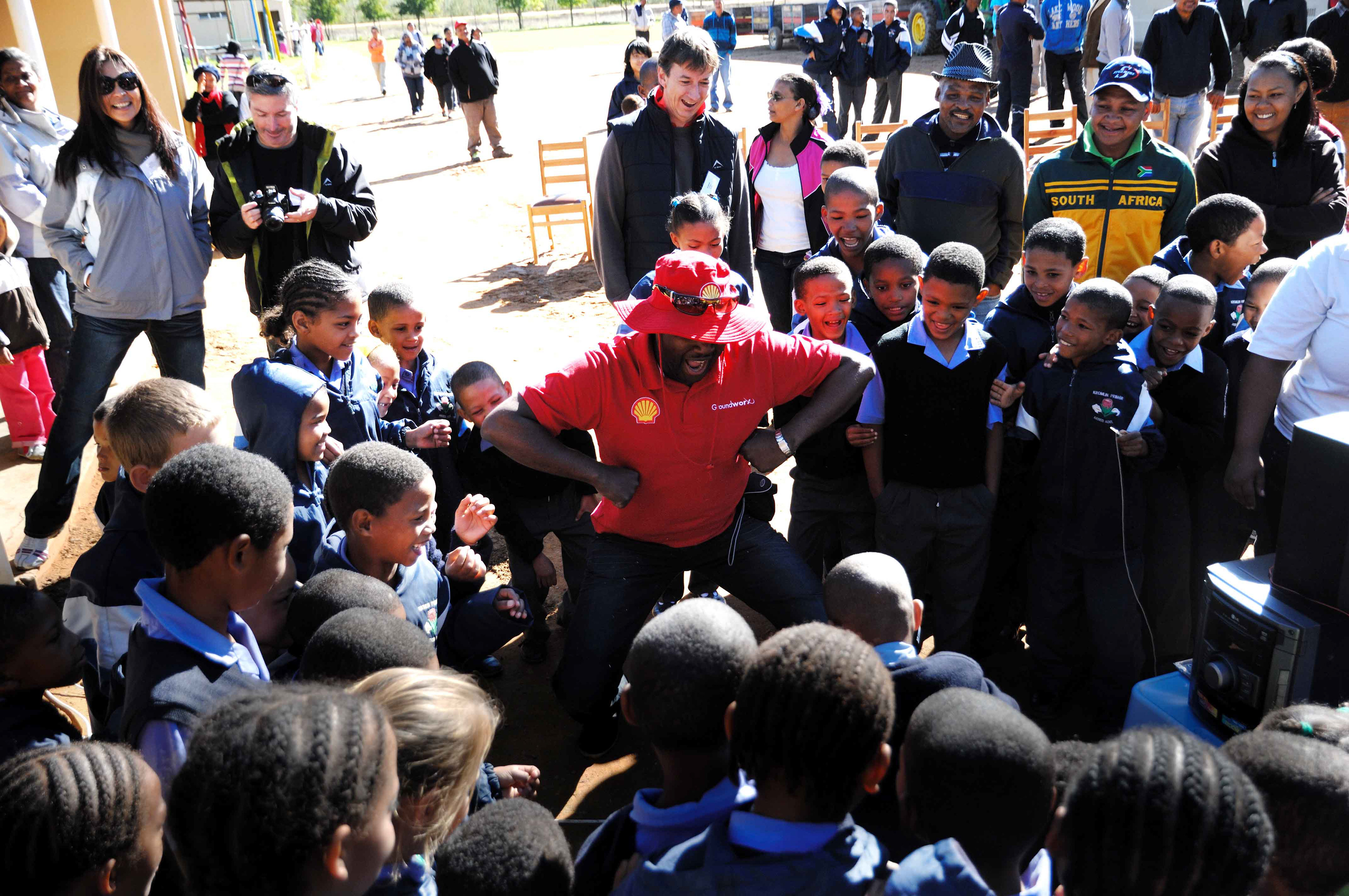 Man in red hat engaging a joyful group of schoolchildren outdoors; onlookers smile, creating a warm, lively atmosphere.